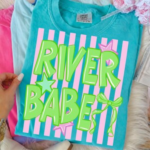 May include: A turquoise Comfort Colors t-shirt with the words "RIVER BABE" in bright green, outlined in white. The text is set against pink vertical stripes. The design includes stars and a bow. The shirt is part of a clothing collection.
