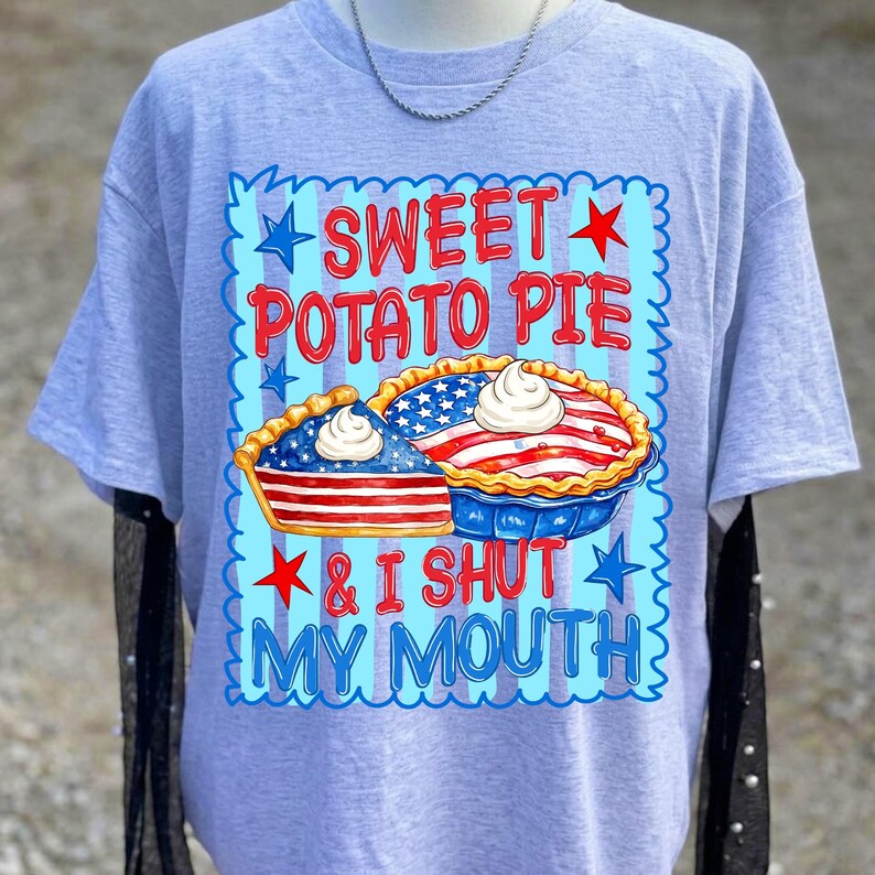 Op de afbeelding: Een lichtgrijs t-shirt met korte mouwen en een grafische print met de tekst SWEET POTATO PIE & I SHUT MY MOUTH. Het ontwerp bevat twee taarten met Amerikaanse vlagpatronen, slagroomtoppings en rode en blauwe sterren op een lichtblauwe achtergrond.