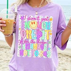 Pode incluir: Uma t-shirt roxo-claro com uma estampa gr&aacute;fica contendo o texto "I don't get ENOUGH CREDIT for the things I MANAGE NOT TO SAY". O design inclui um rosto sorridente amarelo, padr&otilde;es xadrez coloridos, la&ccedil;os cor-de-rosa e pequenas flores.