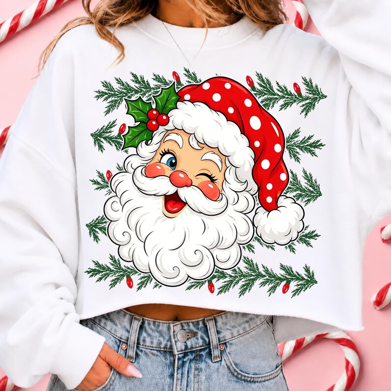May include: A white long-sleeve cropped sweatshirt featuring a graphic print of a winking Santa Claus wearing a red hat with white polka dots. The design includes green pine branches and small red Christmas lights surrounding the Santa face.