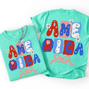 May include: A mint green t-shirt featuring the word AMERICA in a bold, retro font. The letters are filled with red, blue, and white patterns, including stars and small white flowers. A light pink ribbon graphic is positioned below the text.