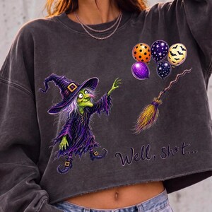 K&ouml;nnte beinhalten: Ein anthrazitfarbenes, bauchfreies Sweatshirt mit einer Grafik einer gr&uuml;nh&auml;utigen Hexe in violettem Gewand und Hut. Daneben befinden sich vier bunte Luftballons und ein Besen. Der Text "Well, sh*t..." ist in violetter Schreibschrift aufgedruckt.