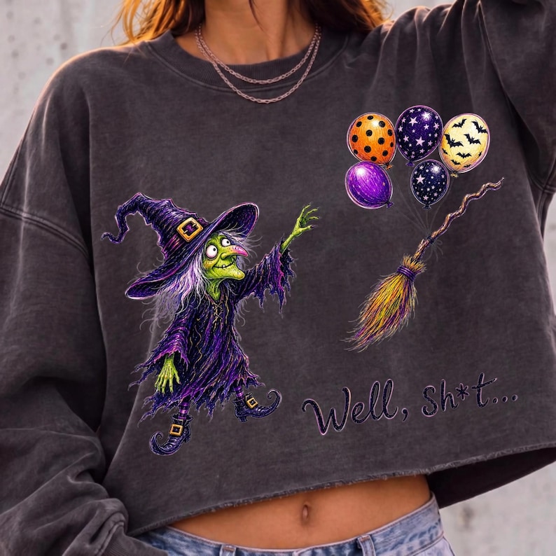 K&ouml;nnte beinhalten: Ein anthrazitfarbenes, bauchfreies Sweatshirt mit einer Grafik einer gr&uuml;nh&auml;utigen Hexe in violettem Gewand und Hut. Daneben befinden sich vier bunte Luftballons und ein Besen. Der Text "Well, sh*t..." ist in violetter Schreibschrift aufgedruckt.