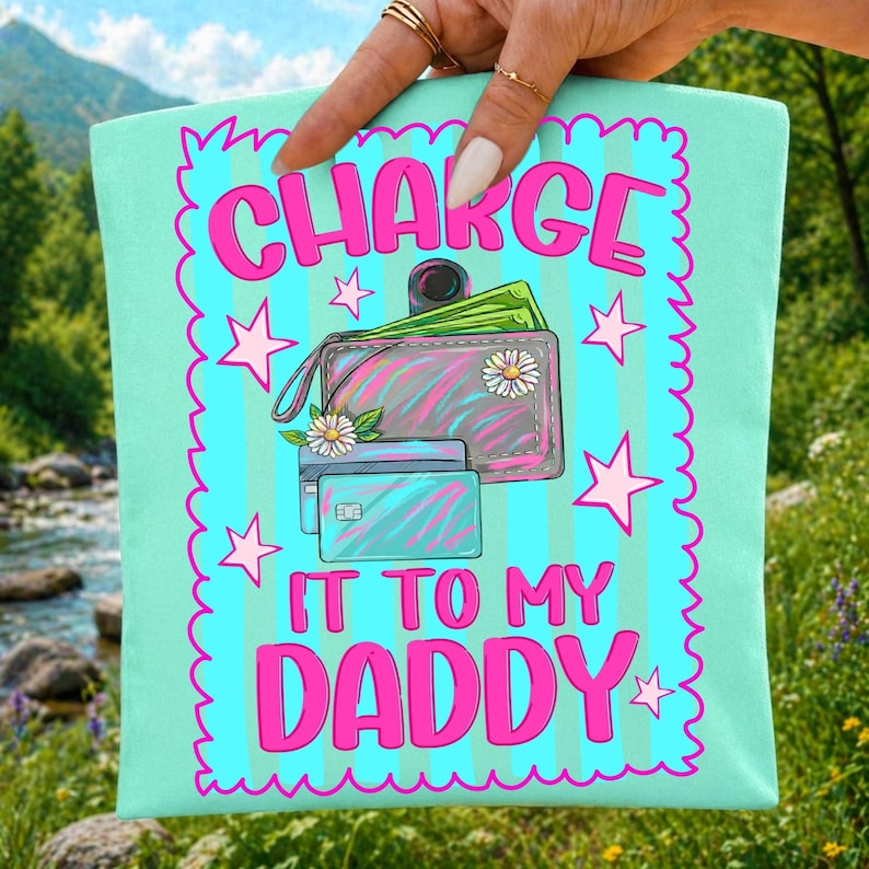 May include: A light mint green tote bag featuring a bright blue rectangular graphic with a scalloped pink border. The design displays a pink wallet with a daisy, two credit cards, and pink stars, with the text CHARGE IT TO MY DADDY in pink.