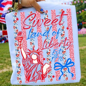 May include: Light blue t-shirt with a patriotic design. The shirt features the Statue of Liberty, red and blue stripes, floral accents, and the text "Sweet Land of Liberty Est. 1776".