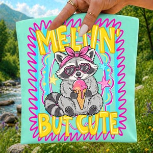 Puede incluir: Un cuadrado de tela verde menta claro con un mapache de dibujos animados que lleva una diadema rosa y gafas de sol en forma de coraz&oacute;n. El mapache sostiene un helado derriti&eacute;ndose. El texto dice MELTIN' BUT CUTE en letras amarillas con un borde rosa.