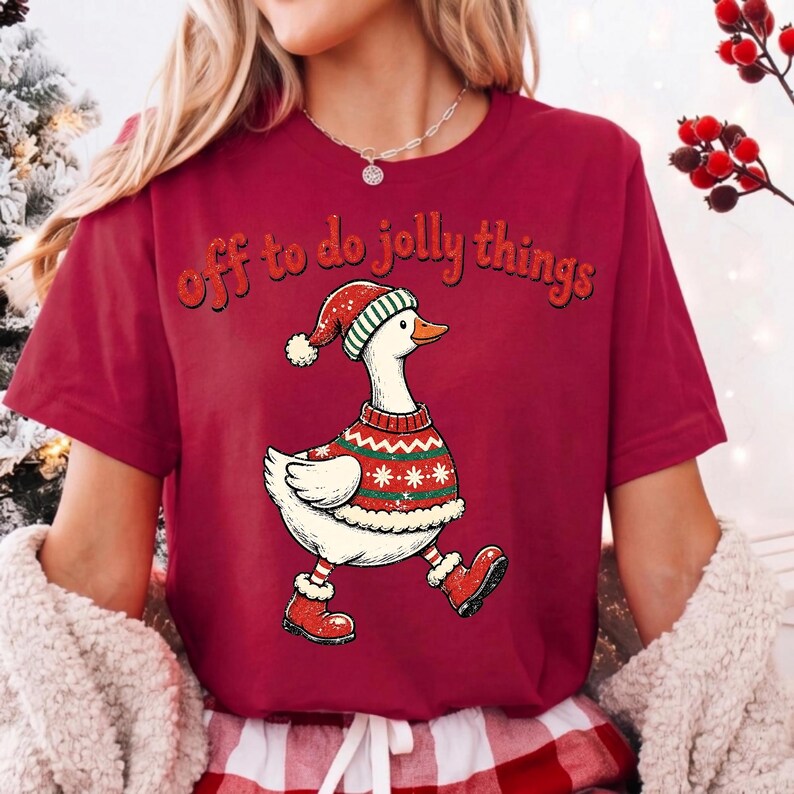 May include: A maroon short-sleeved t-shirt featuring a graphic of a white goose wearing a red and green patterned sweater, a matching Santa hat, and red boots. The text "off to do jolly things" is printed in a dark red font above the goose.