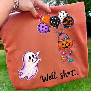 May include: A rust-colored fabric item featuring a white ghost graphic, a pumpkin-shaped basket filled with colorful candies, and a cluster of patterned balloons. The text "Well, sh*t..." is printed in black script at the bottom right corner.