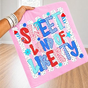 K&ouml;nnte beinhalten: Ein quadratischer rosa Stoffartikel mit dem Text SWEET LAND OF LIBERTY in einer Mischung aus rot-wei&szlig;-blauen Karomustern und Punkten. Das Design enth&auml;lt kleine Feuerwerksillustrationen und blaue Blumen auf einem wei&szlig; gestreiften Hintergrund.