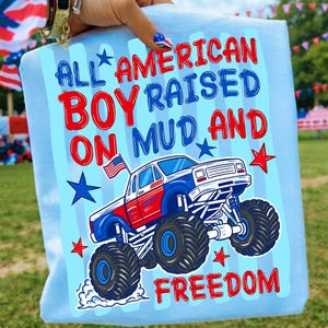 May include: A light blue t-shirt featuring a graphic of a red, white, and blue monster truck with a small American flag. The text reads ALL AMERICAN BOY RAISED ON MUD AND FREEDOM, accompanied by red and blue stars on a light blue striped background.