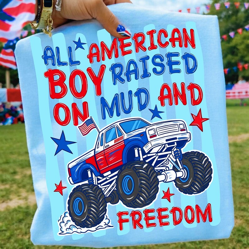 May include: A light blue t-shirt featuring a graphic of a red, white, and blue monster truck with a small American flag. The text reads ALL AMERICAN BOY RAISED ON MUD AND FREEDOM, accompanied by red and blue stars on a light blue striped background.