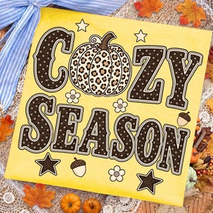 Może przedstawiać: Kwadratowy ż&oacute;łty znak z napisem COZY SEASON dużymi ciemnobrązowymi literami w białe kropki. Pomiędzy słowami znajduje się dynia w panterkę. Wz&oacute;r zawiera małe białe kwiatki, gwiazdki oraz żołędzie.