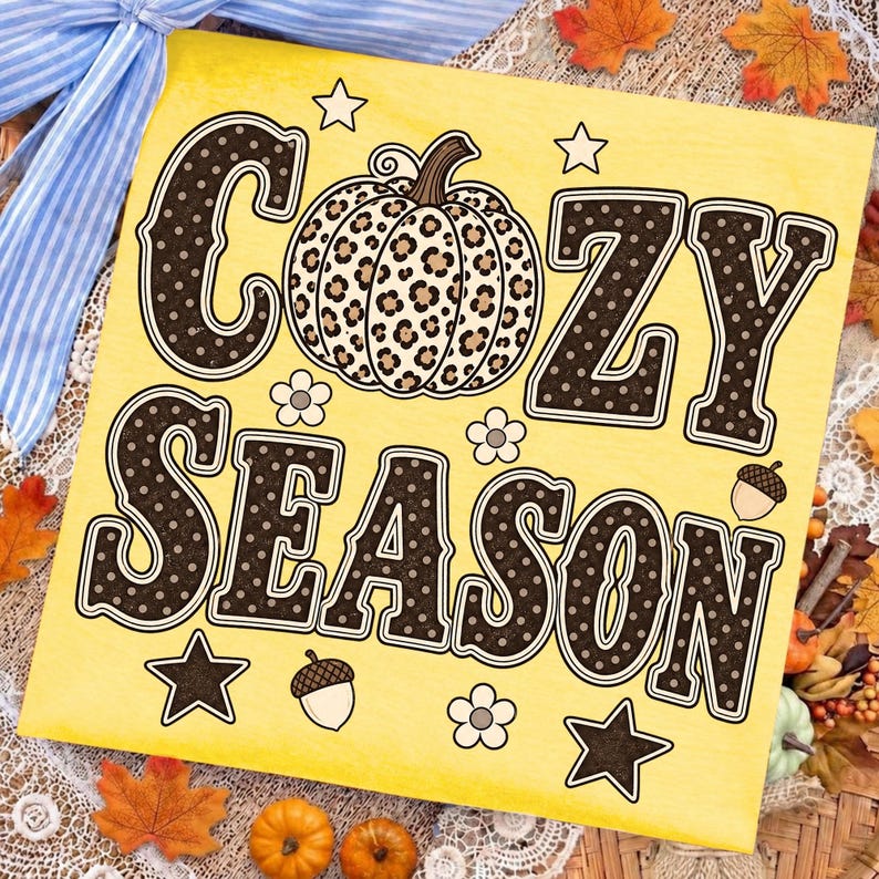 Może przedstawiać: Kwadratowy ż&oacute;łty znak z napisem COZY SEASON dużymi ciemnobrązowymi literami w białe kropki. Pomiędzy słowami znajduje się dynia w panterkę. Wz&oacute;r zawiera małe białe kwiatki, gwiazdki oraz żołędzie.