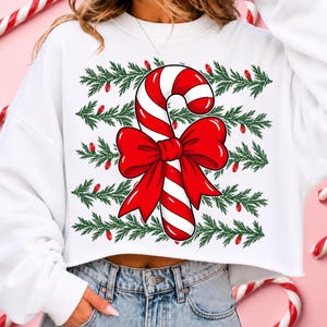 May include: A white cropped sweatshirt featuring a graphic print of a red and white striped candy cane tied with a large red bow. The design is layered over horizontal rows of green pine branches interspersed with small red holiday light bulbs.