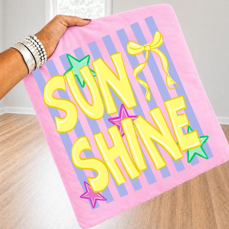 May include: A pink blanket with the words "SUN SHINE" in yellow, outlined in white. The blanket has blue and white stripes, yellow bows, and green and purple star accents. The blanket is held up by a person.