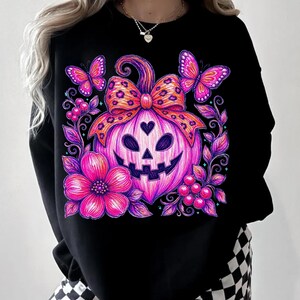 K&ouml;nnte beinhalten: Ein schwarzes Langarm-Sweatshirt mit einem lebendigen Grafikdruck eines pinken und violetten K&uuml;rbis-Gesichts mit herzf&ouml;rmiger Nase. Der K&uuml;rbis ist mit einer Schleife im Leopardenmuster, zwei Schmetterlingen, einer Blume und Beeren verziert.