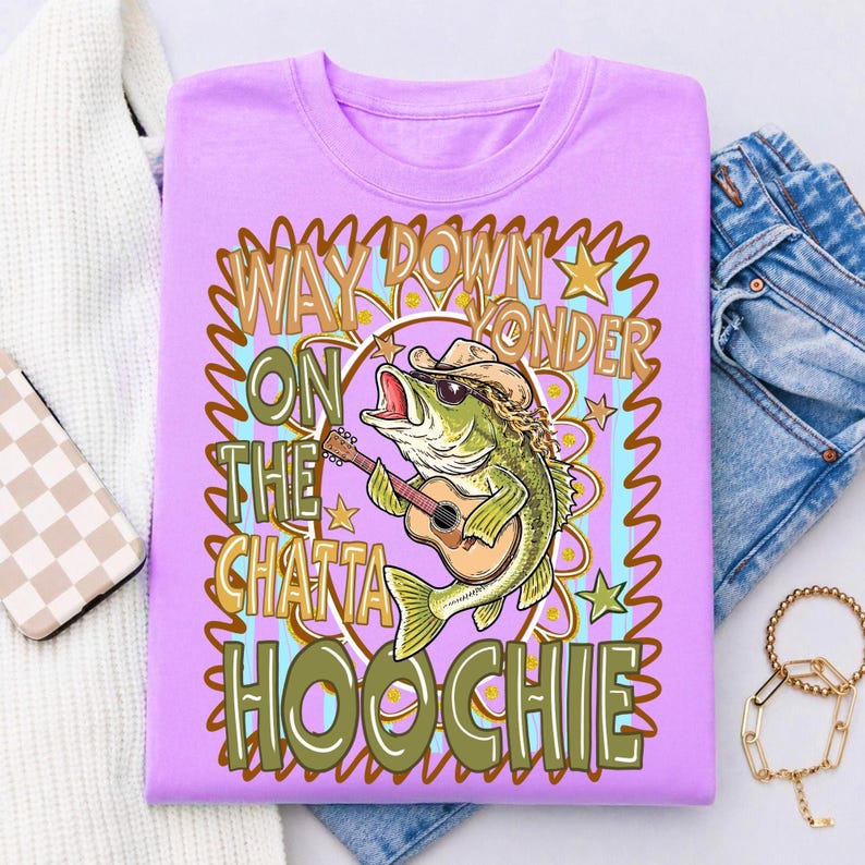 May include: A lilac-colored t-shirt features a cartoon bass fish wearing a cowboy hat and sunglasses, playing a guitar. The text reads "Way Down Yonder On The Chatta Hoochie" in a decorative font, with stars and a colorful border.