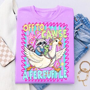 May include: A lilac-colored t-shirt featuring a whimsical illustration. A raccoon wearing overalls and sunglasses rides on a goose, accompanied by a frog. The text reads "OFF TO CAWSE A FERFUFFLE" in a playful font, surrounded by stars and wavy lines.