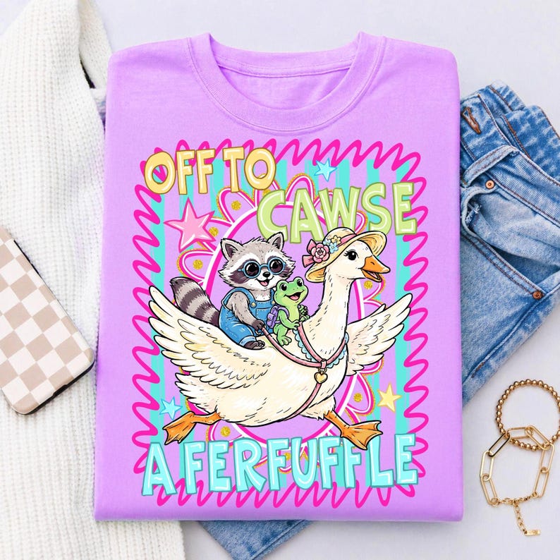 May include: A lilac-colored t-shirt featuring a whimsical illustration. A raccoon wearing overalls and sunglasses rides on a goose, accompanied by a frog. The text reads "OFF TO CAWSE A FERFUFFLE" in a playful font, surrounded by stars and wavy lines.
