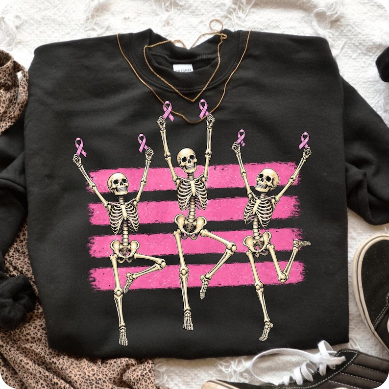 May include: A black crewneck sweatshirt featuring a graphic of three beige skeletons dancing in front of horizontal pink brushstrokes. Each skeleton holds a small pink ribbon above its head. A thin gold-toned chain necklace rests at the neckline.