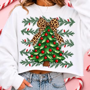 Op de afbeelding: Een wit sweatshirt met lange mouwen en een afbeelding van een groene kerstboom versierd met rode kerstballen en een grote strik met luipaardprint. De achtergrond van de afbeelding bevat horizontale rijen groene dennentakken en rode lichtjes.