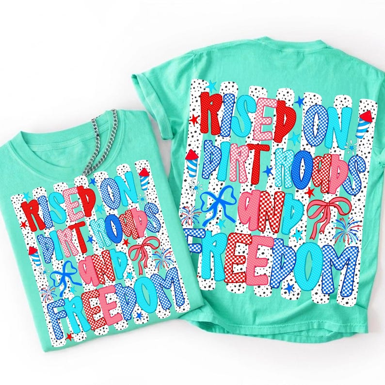 May include: Two mint green t-shirts featuring a graphic print with the text "RISED ON DIRT ROADS AND FREEDOM". The design includes red, white, and blue patterns, stars, fireworks, and bows over vertical white stripes with black polka dots.