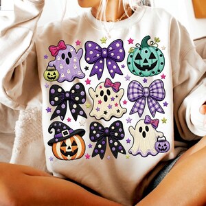 K&ouml;nnte beinhalten: Ein beigefarbenes Sweatshirt mit einem Grafikdruck von drei Geistern, drei K&uuml;rbissen und drei Schleifen. Die Geister sind wei&szlig; und lila, die K&uuml;rbisse orange und blaugr&uuml;n, und die Schleifen sind lila, schwarz und kariert, verziert mit kleinen Sternen.