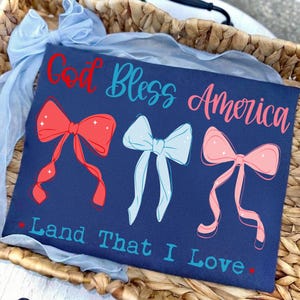 May include: A navy blue t-shirt featuring the red and light blue text God Bless America and Land That I Love. The design includes three illustrated bows in red, light blue, and pink, each accented with small white star details on the fabric.