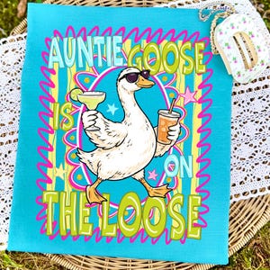 K&ouml;nnte beinhalten: Ein t&uuml;rkisfarbenes K&uuml;chentuch mit einer Cartoon-Gans, die eine Sonnenbrille tr&auml;gt und eine Margarita und ein Getr&auml;nk mit Strohhalm h&auml;lt. Der Text lautet "Auntie Goose is on the Loose" in einem farbenfrohen Design.