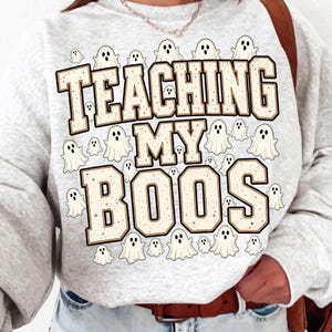 May include: A light gray heathered sweatshirt featuring the text TEACHING MY BOOS in large, blocky, cream-colored letters with dark outlines. The design is surrounded by numerous small, white cartoon ghost illustrations with black eyes.