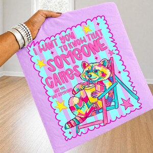 K&ouml;nnte beinhalten: Ein hellviolettes Stoffquadrat mit einem bunten Waschb&auml;ren, der in einem rosa Stuhl sitzt und ein gelbes Getr&auml;nk h&auml;lt. Der Text lautet: I WANT YOU TO KNOW THAT SOMEONE CARES, NOT ME, BUT SOMEONE. Das Design enth&auml;lt gelbe Sterne und einen gewellten Rand.