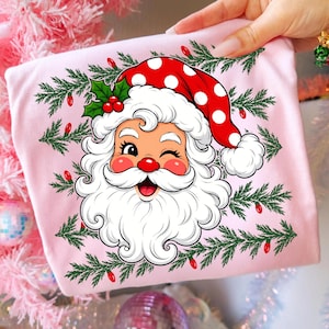 Puede incluir: Una camiseta de color rosa claro con un gr&aacute;fico de un Santa Claus gui&ntilde;ando un ojo y llevando un gorro rojo con lunares blancos. La ilustraci&oacute;n est&aacute; rodeada de ramas de pino verdes y peque&ntilde;as luces navide&ntilde;as rojas, creando un dise&ntilde;o festivo.
