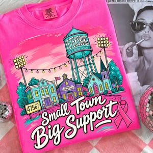 May include: A bright pink t-shirt featuring a graphic illustration of a small town with a water tower labeled PETERSBURG, a city hall, a community school, and a church. The text reads Small Town Big Support with a pink awareness ribbon.