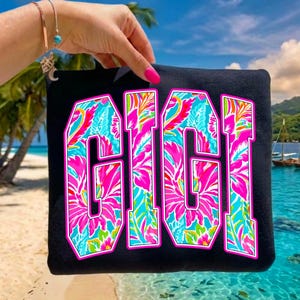 以下が含まれることがあります： 黒色のトートバッグ。中央に「GIGI」の文字が大きくピンク色で白の縁取りで描かれています。文字はピンク、青、オレンジ、緑の色合いのカラフルな花柄で埋められています。背景には熱帯のビーチの風景が写っています。