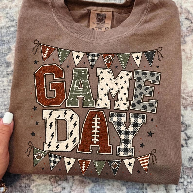 May include: A brown short-sleeved t-shirt featuring a graphic print with the text GAME DAY. The letters are decorated with football-themed patterns including a field, stars, helmets, lightning bolts, and a football. Bunting flags border the text.