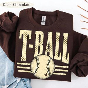 Pode incluir: Uma camisola de gola redonda em castanho chocolate escuro com o texto T-BALL em letras grandes de cor creme com bolinhas. Abaixo do texto, h&aacute; um gr&aacute;fico de uma bola de basebol com um pequeno cora&ccedil;&atilde;o, centrado sobre tr&ecirc;s linhas.