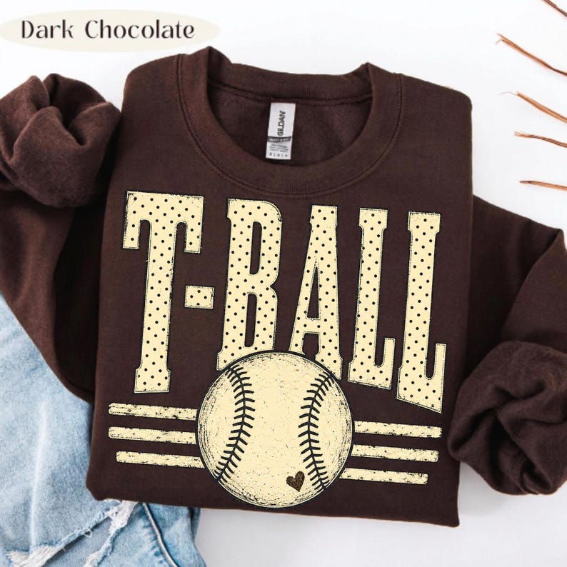 Pode incluir: Uma camisola de gola redonda em castanho chocolate escuro com o texto T-BALL em letras grandes de cor creme com bolinhas. Abaixo do texto, h&aacute; um gr&aacute;fico de uma bola de basebol com um pequeno cora&ccedil;&atilde;o, centrado sobre tr&ecirc;s linhas.