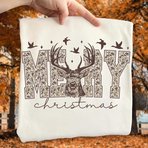 May include: Cream-colored t-shirt featuring the text "MERRY CHRISTMAS" with a camouflage pattern. The letter design includes a detailed illustration of a deer head with antlers. Above the text are flying birds and diamond shapes.