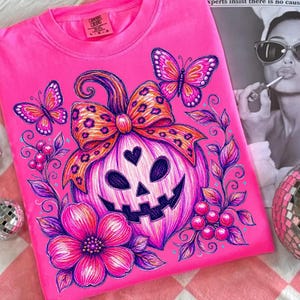 K&ouml;nnte beinhalten: Ein leuchtend pinkes T-Shirt mit einer Grafik eines K&uuml;rbisses mit herzf&ouml;rmiger Nase, der mit einer Schleife im Leopardenmuster verziert ist. Das Design zeigt zwei Schmetterlinge, eine gro&szlig;e Blume und Beeren in Pink-, Lila- und Oranget&ouml;nen.