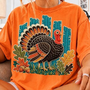 May include: An orange short-sleeve t-shirt featuring a large graphic of a turkey with black, brown, and orange feathers. The design includes a teal vertical striped background, green and orange oak leaves, and two brown acorns at the bottom.