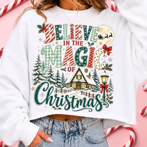 May include: A white cropped sweatshirt featuring a colorful graphic with the text "BELIEVE IN THE MAGIC OF CHRISTMAS". The design includes a snowy cabin, pine trees, a street lamp, a moon with a sleigh silhouette, and festive red and green patterns.