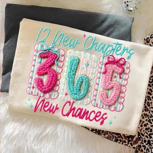 Puede incluir: Camiseta color crema con texto y n&uacute;meros bordados. El texto dice "12 New Chapters" y "365 New Chances" en turquesa y rosa. Los n&uacute;meros "3", "6" y "5" son en rosa y turquesa, con bordes decorativos.