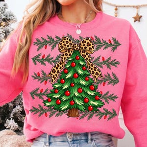 Op de afbeelding: Een felroze sweatshirt met een afbeelding van een groene kerstboom versierd met rode kerstballen en bekroond met een grote strik met luipaardprint. De achtergrond van het ontwerp bevat horizontale dennentakken en kleine rode lampjes.