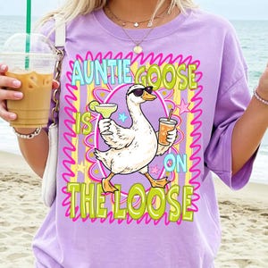 K&ouml;nnte beinhalten: Ein lila T-Shirt mit einer Grafik einer Gans mit Sonnenbrille, die einen Cocktail und ein Getr&auml;nk h&auml;lt. Der Text lautet "AUNTIE GOOSE IS ON THE LOOSE" in Gr&uuml;n und Gelb.