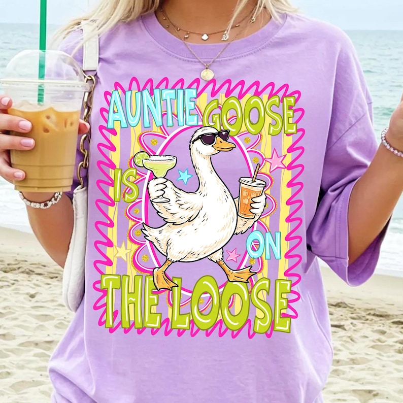 K&ouml;nnte beinhalten: Ein lila T-Shirt mit einer Grafik einer Gans mit Sonnenbrille, die einen Cocktail und ein Getr&auml;nk h&auml;lt. Der Text lautet "AUNTIE GOOSE IS ON THE LOOSE" in Gr&uuml;n und Gelb.