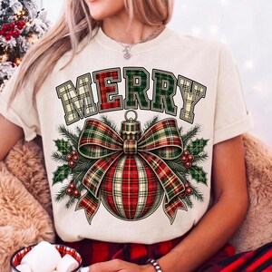 Puede incluir: Una camiseta de color crema con la palabra MERRY en una fuente de cuadros rojos y verdes. Debajo del texto hay un gr&aacute;fico de un adorno navide&ntilde;o grande decorado con un lazo de cuadros a juego, ramas de pino verdes y bayas de acebo rojas.