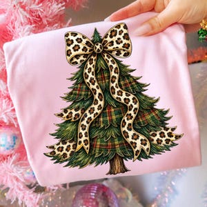 May include: A light pink t-shirt featuring a graphic illustration of a green Christmas tree with a red, green, and gold plaid pattern. The tree is adorned with a large, flowing bow featuring a tan and black leopard print pattern.