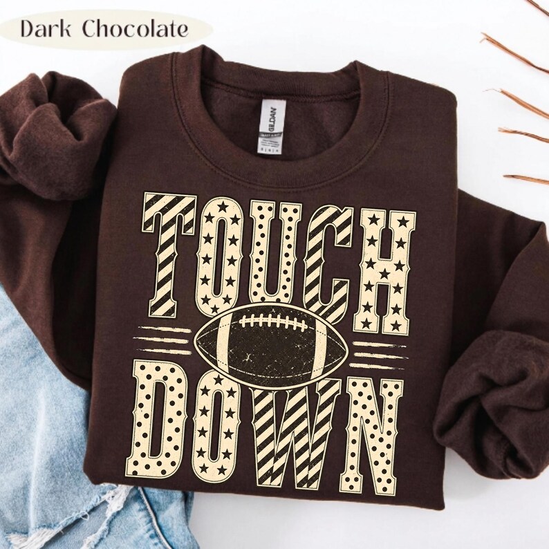 以下が含まれることがあります： ダークチョコレートブラウンのクルーネックトレーナー。クリーム色のフォントで「TOUCH DOWN」と書かれており、星、ストライプ、水玉模様があしらわれています。「TOUCH」と「DOWN」の文字の間には、アメリカンフットボールのグラフィックが配置されています。