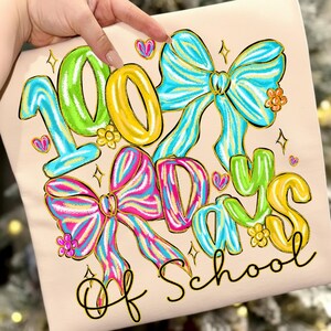 Op de afbeelding: Lichtbeige T-shirt met de tekst "100 Days of School" in kleurrijke letters. Het ontwerp bevat grote strikken, harten en bloemen in blauw, roze, geel en groen, perfect voor een speciale schooldag.
