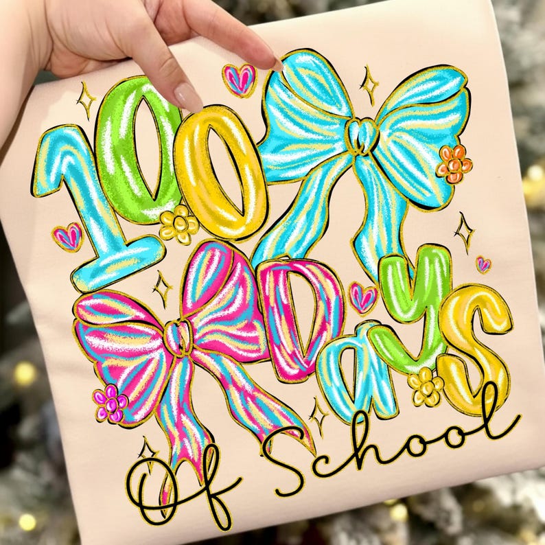 Op de afbeelding: Lichtbeige T-shirt met de tekst "100 Days of School" in kleurrijke letters. Het ontwerp bevat grote strikken, harten en bloemen in blauw, roze, geel en groen, perfect voor een speciale schooldag.